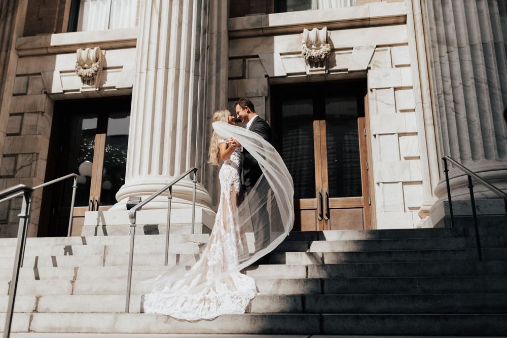Wedding couple embraces on historic building steps with flowing veil caught in motion.