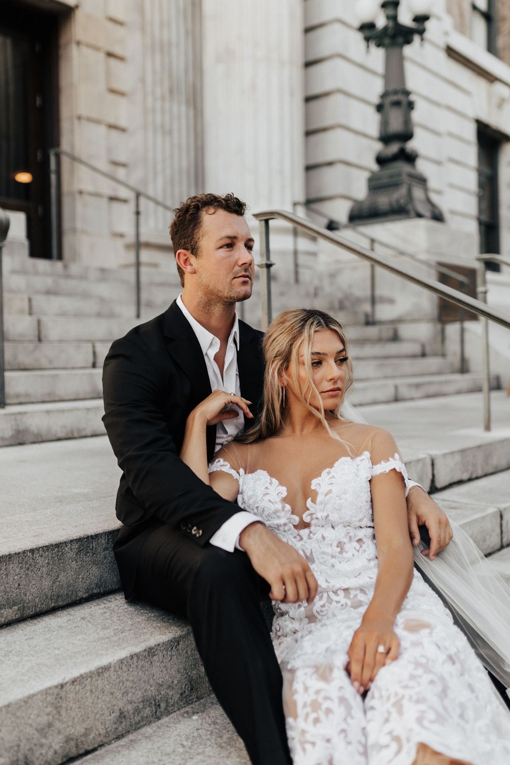 A romantic wedding couple sits on stone steps outside a grand building in elegant attire.