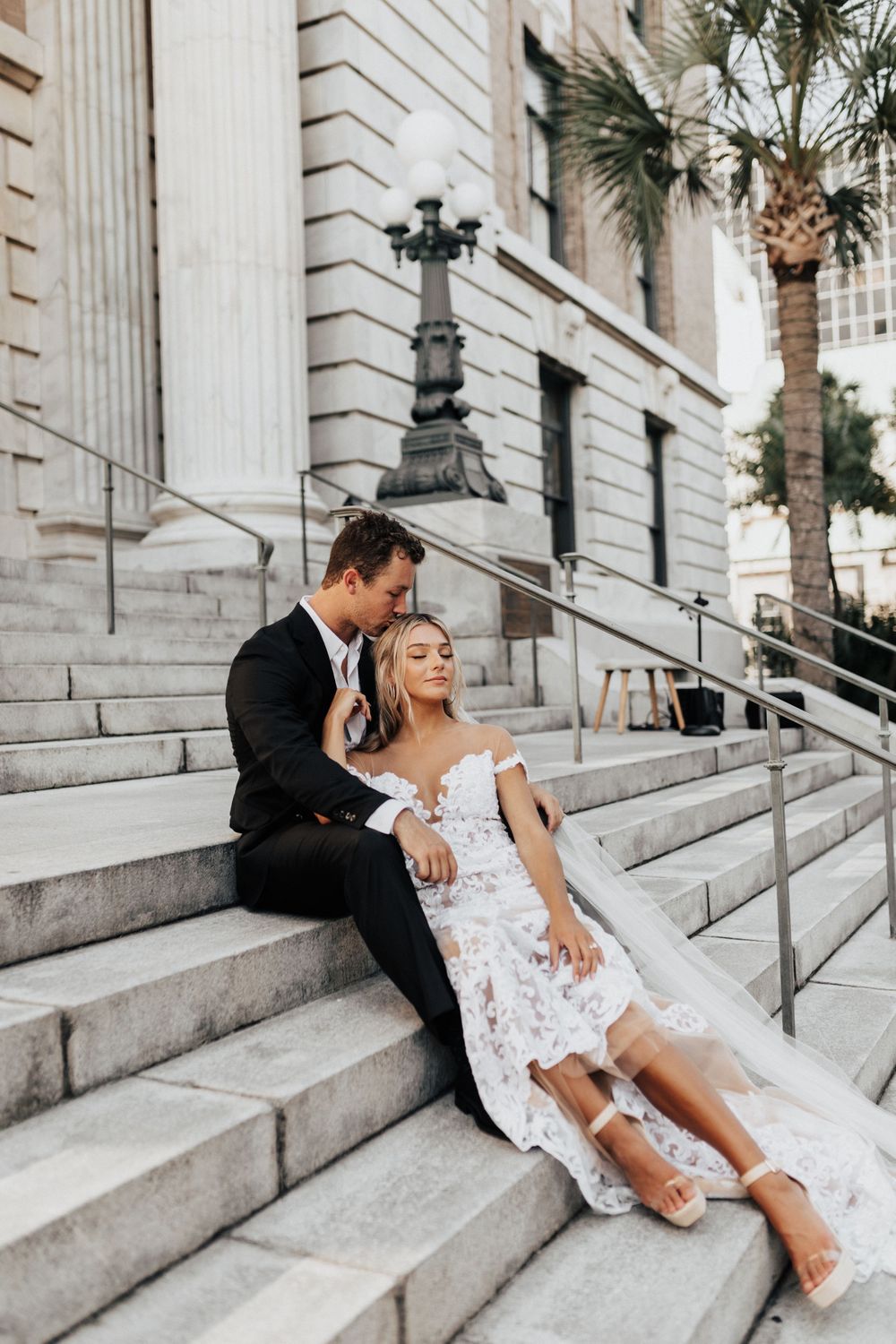 Wedding couple shares intimate moment on stone steps with palm tree in background.