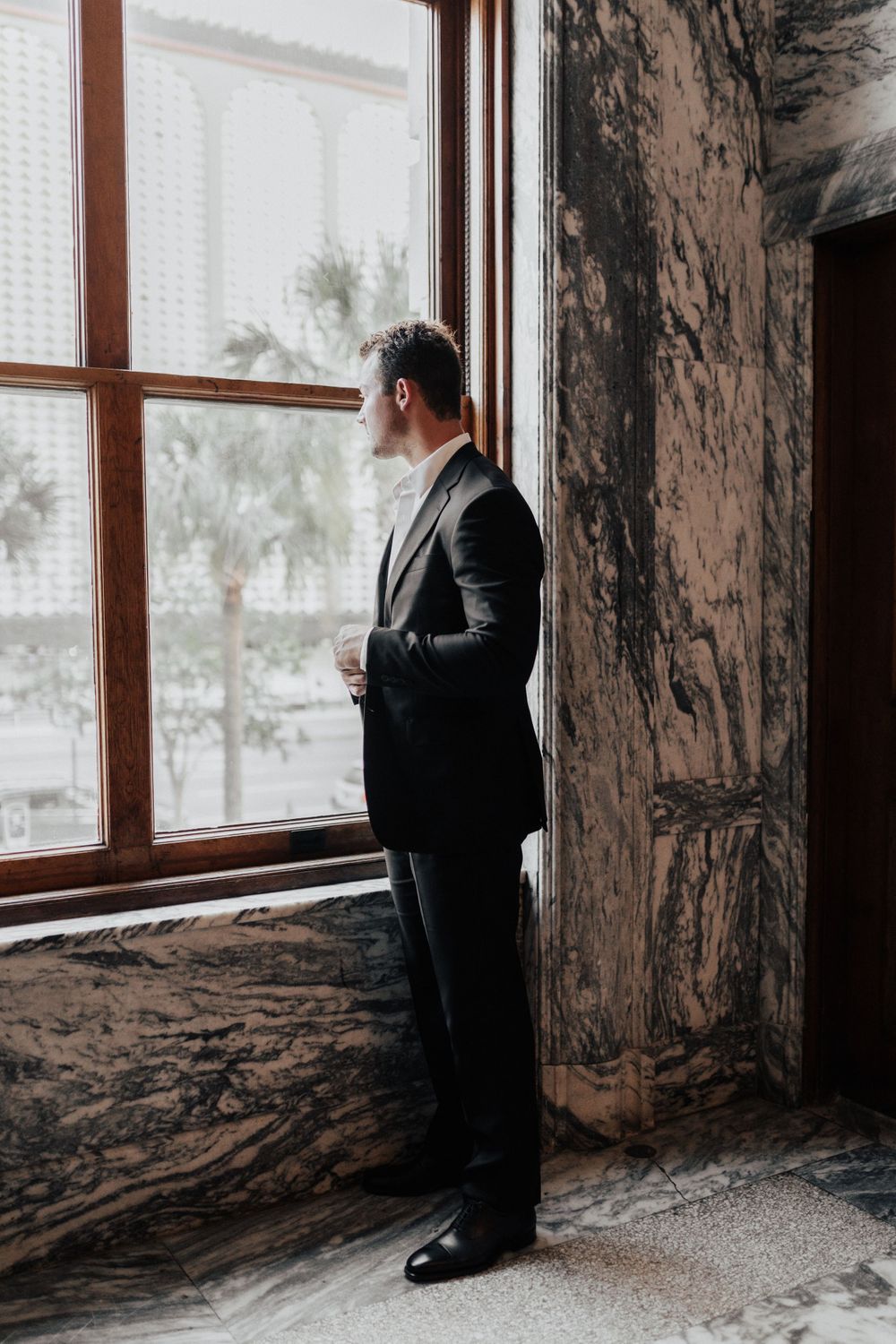 A contemplative moment by a large window featuring a groom in a classic black suit.