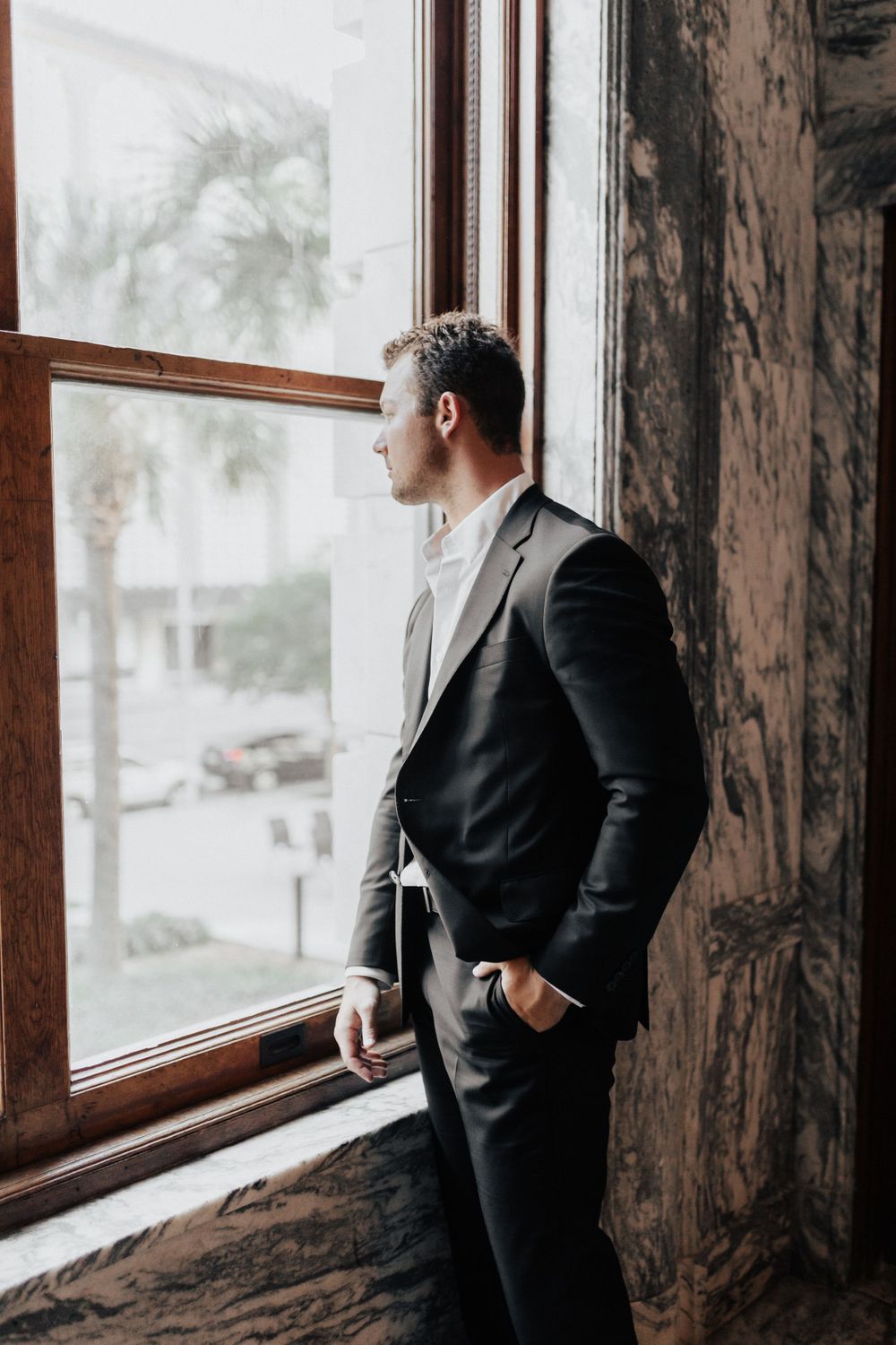 A person in a dark suit gazes thoughtfully through a large window in an elegant room with marble walls.