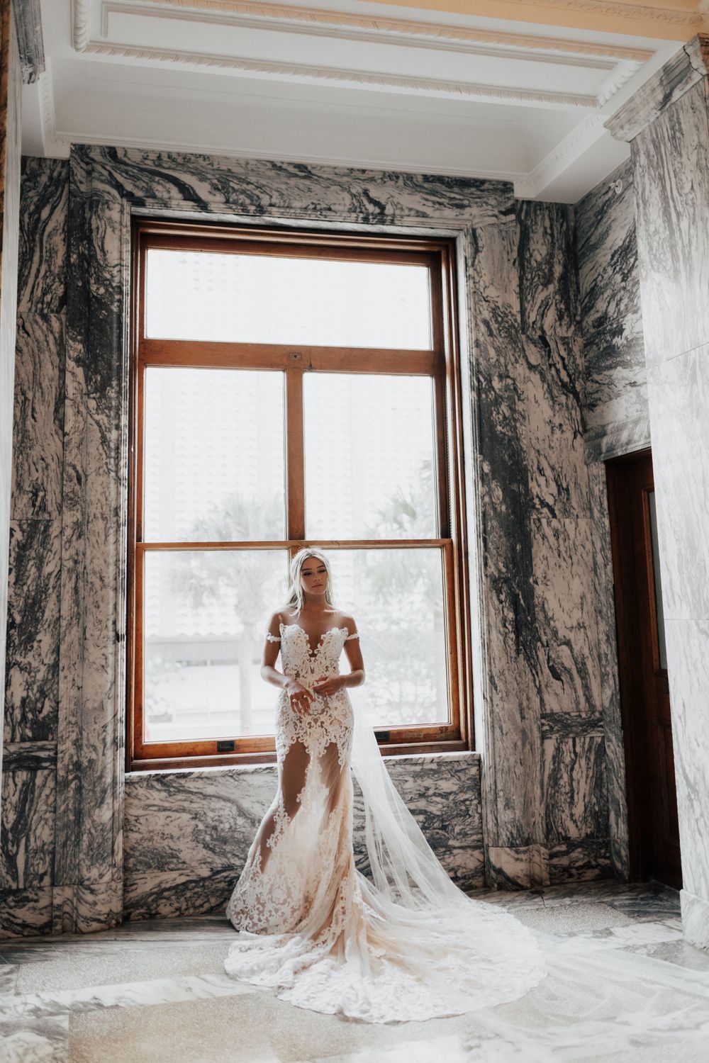 A stunning lace wedding gown with flowing train photographed in front of a tall window in a marble room.