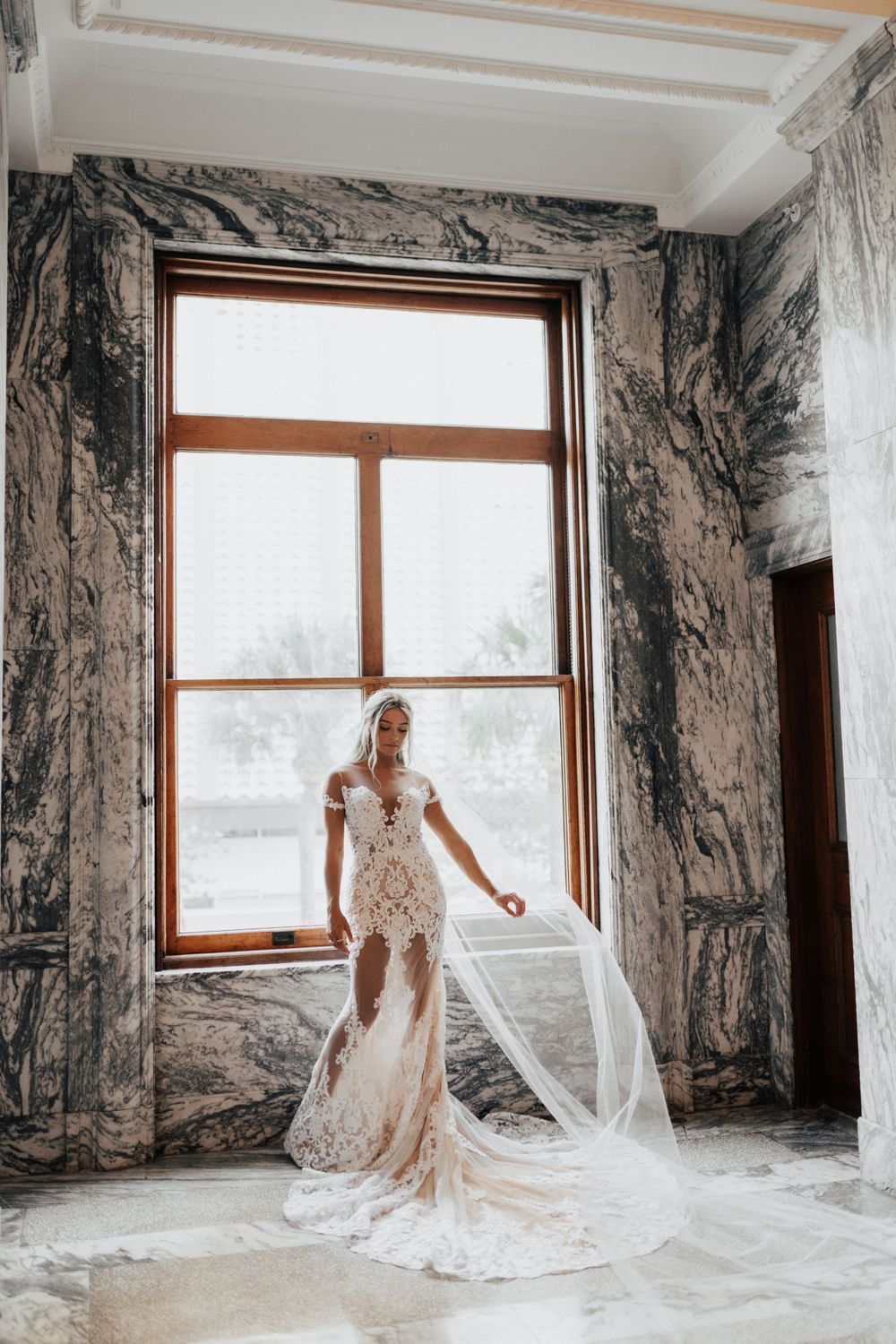 An elegant bride poses in a sheer lace wedding dress with long train against marble walls and window.