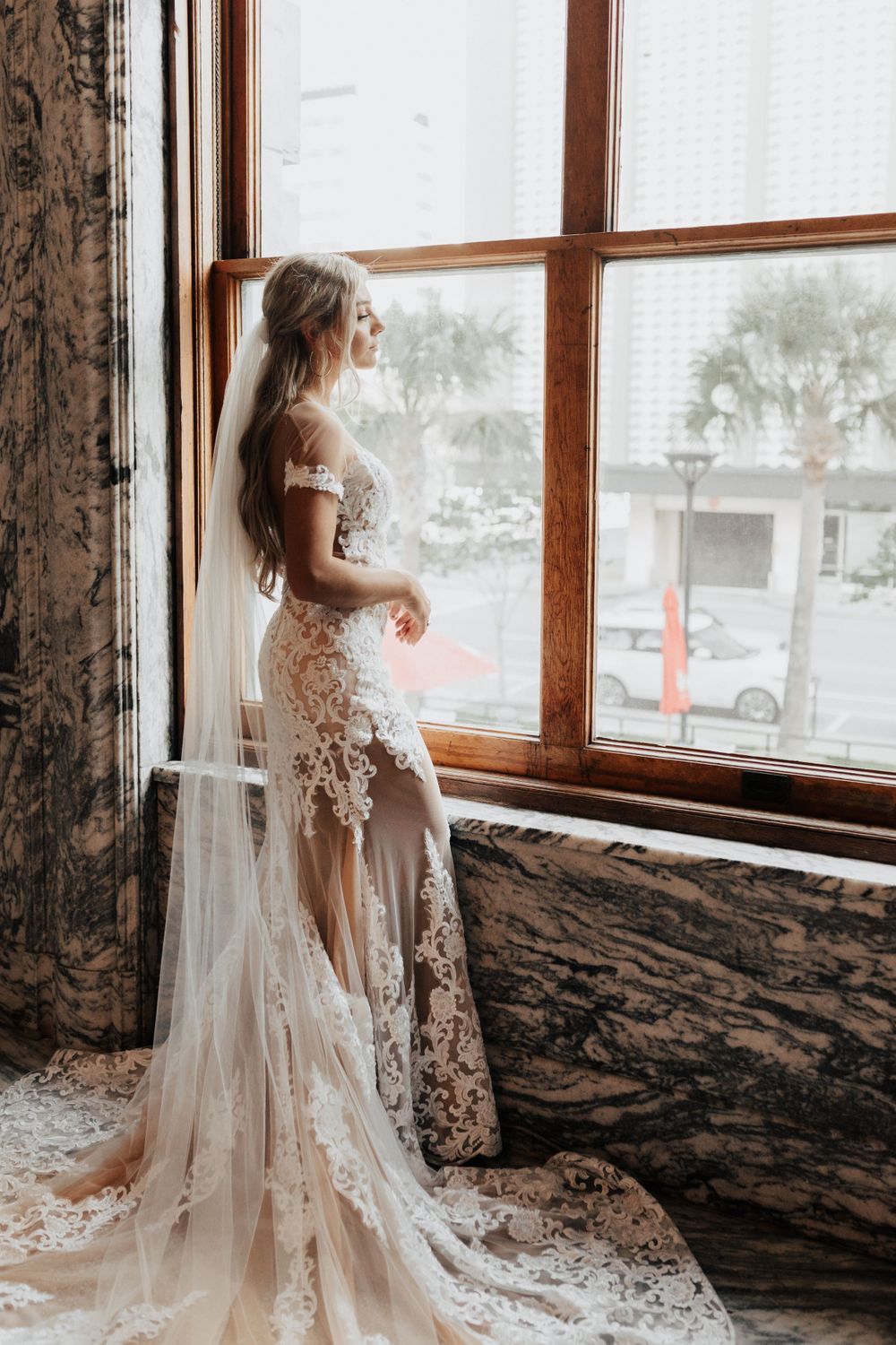 A delicate lace wedding dress with flowing train captured in front of a bright window with marble surroundings.