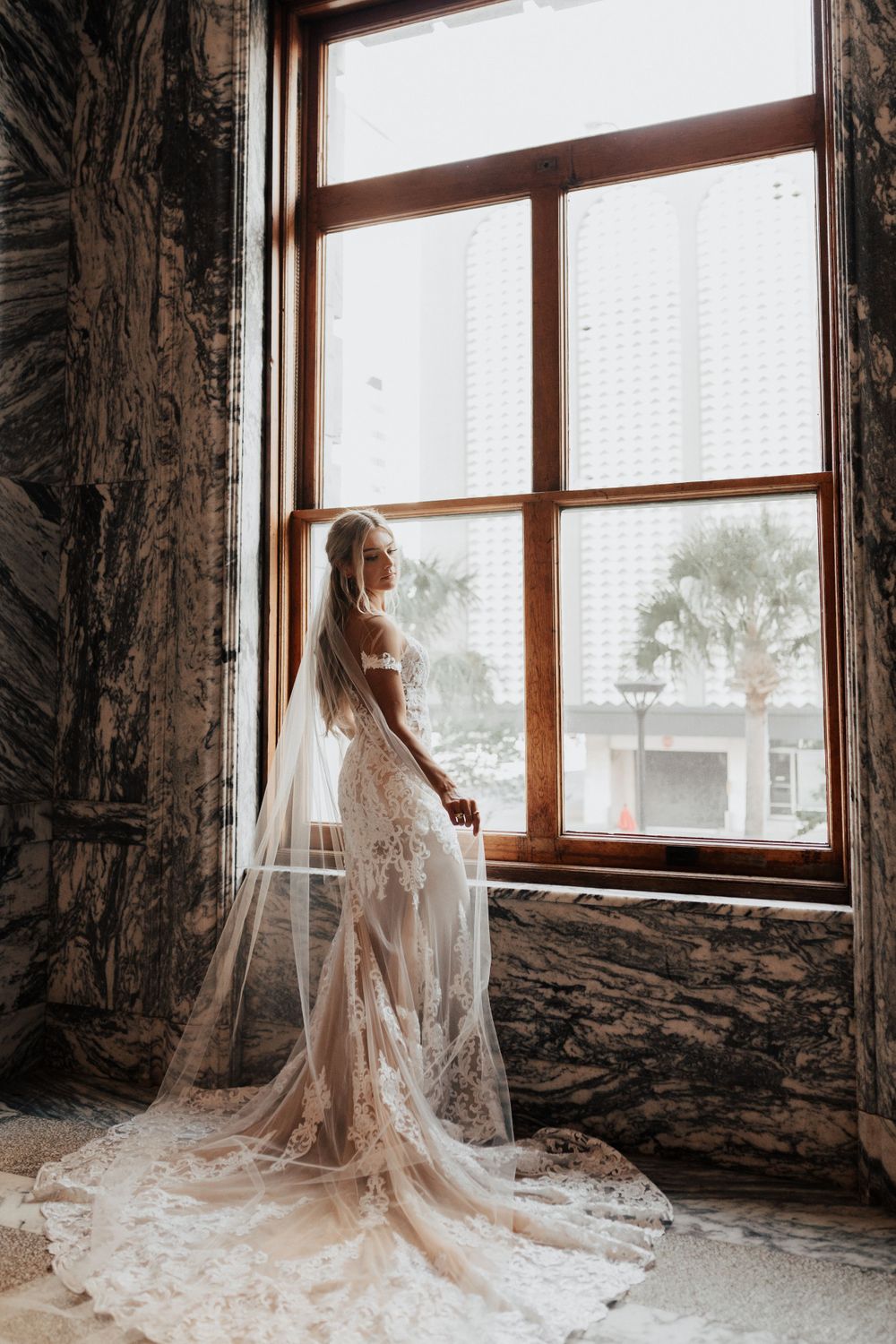 An elegant wedding gown with sheer details and long train silhouetted against a bright window.