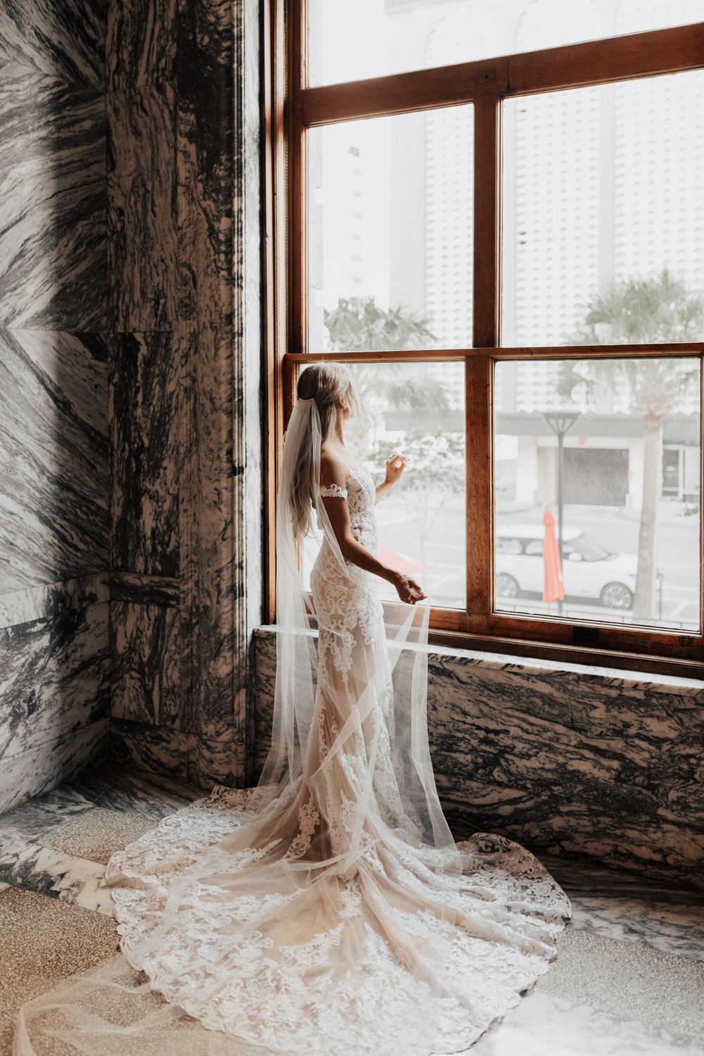 A wedding dress with intricate lace details and flowing train photographed by a large window.