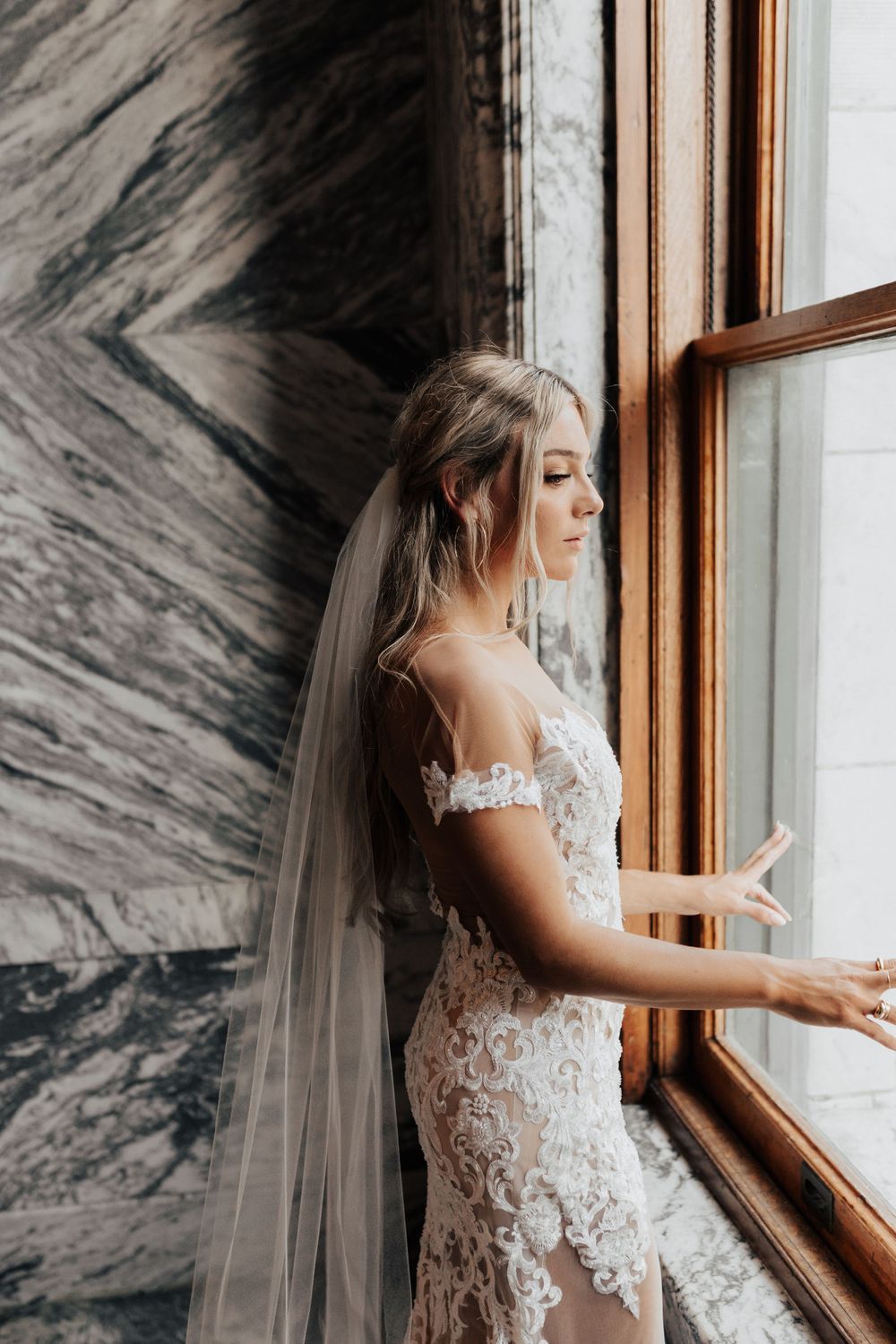 A delicate off-shoulder wedding dress photographed against dramatic marble walls and window.