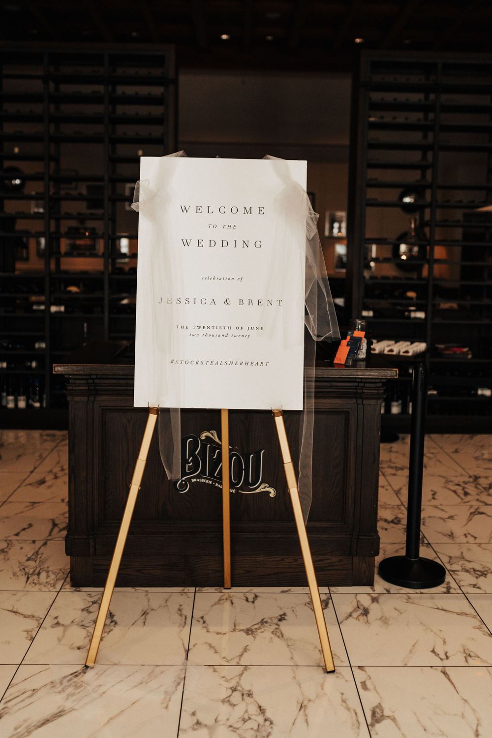 A minimalist white wedding welcome sign displayed on a gold easel against marble flooring.