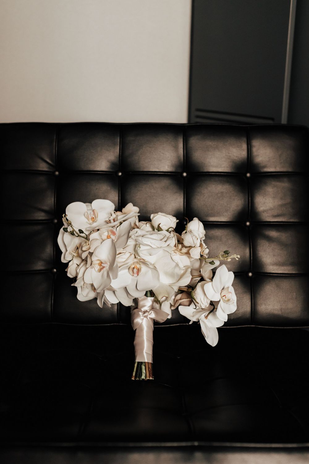 An elegant white orchid and rose wedding bouquet rests on a black leather tufted couch.