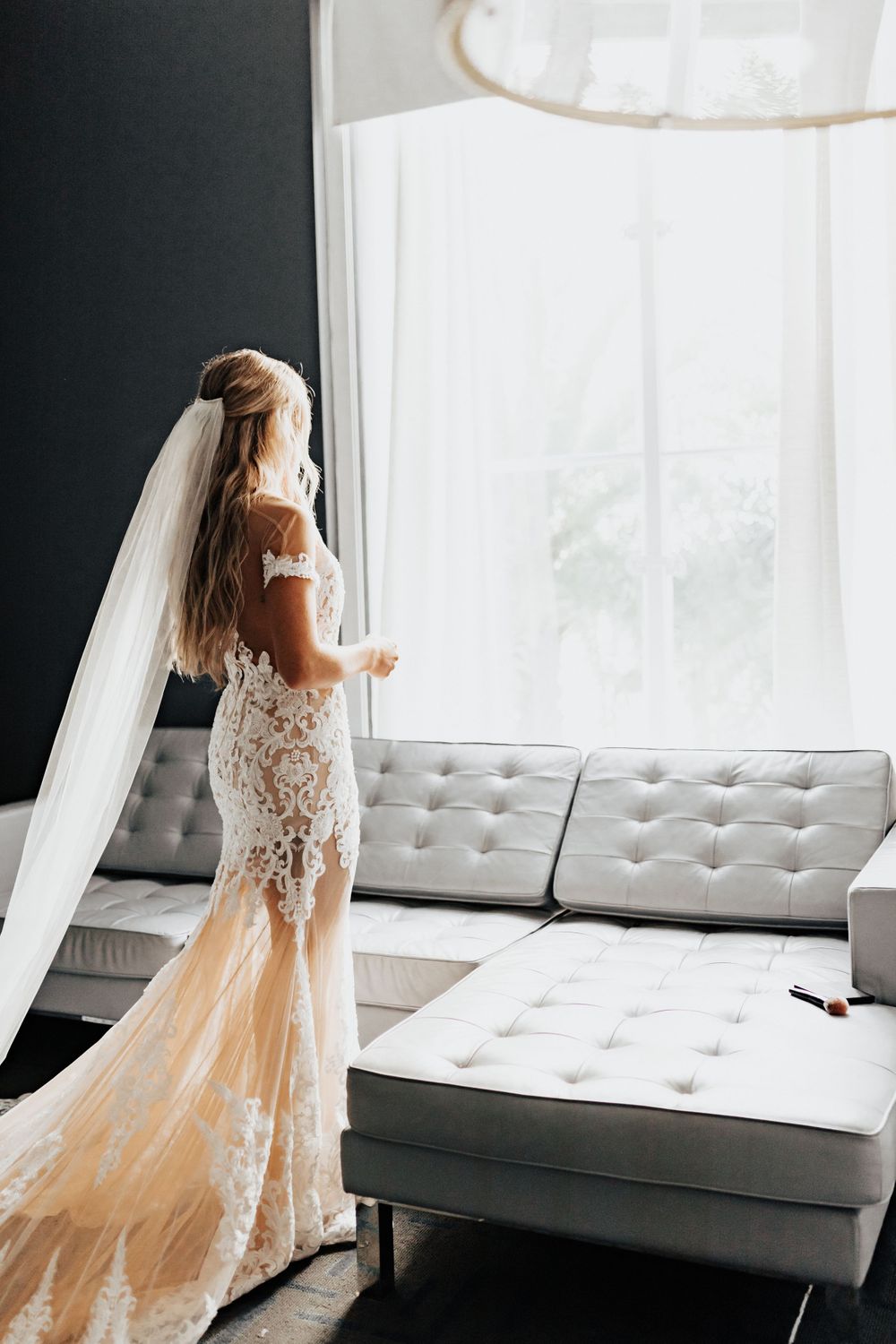 A stunning lace wedding dress with flowing veil stands elegantly by a bright window.