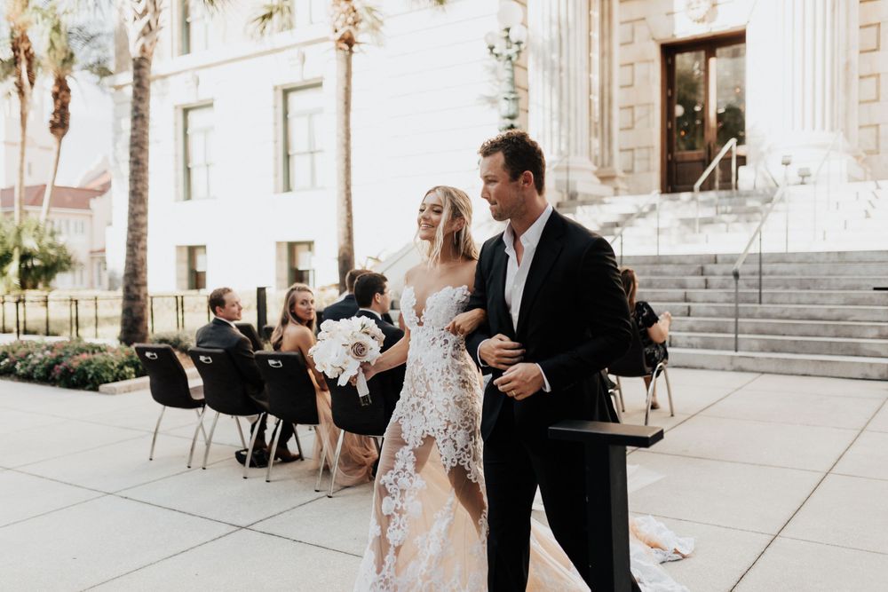 A newlywed couple walks along a sunny path outside a classical building after their ceremony.