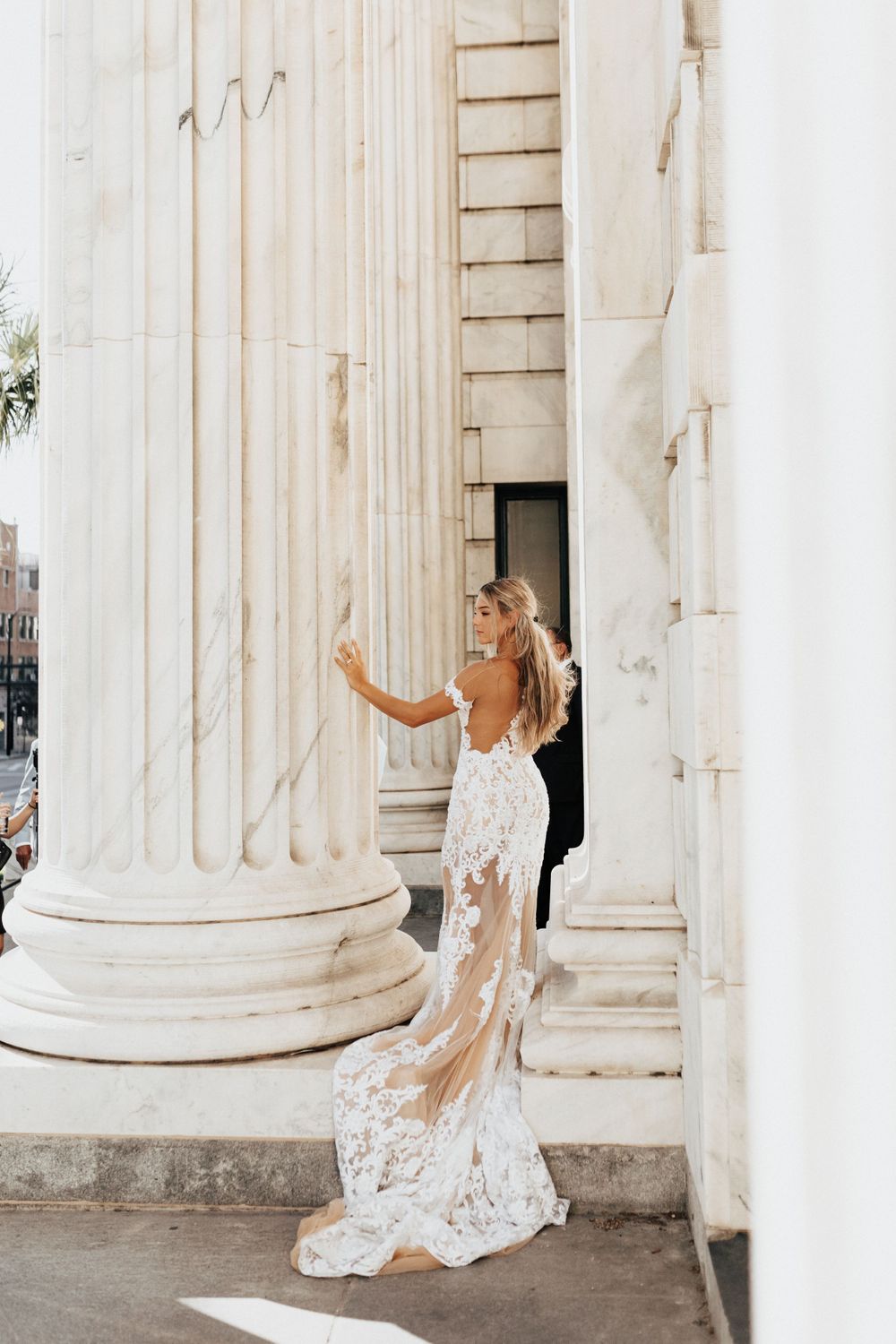 A bride in a lace gown poses against white marble columns of a historic building.