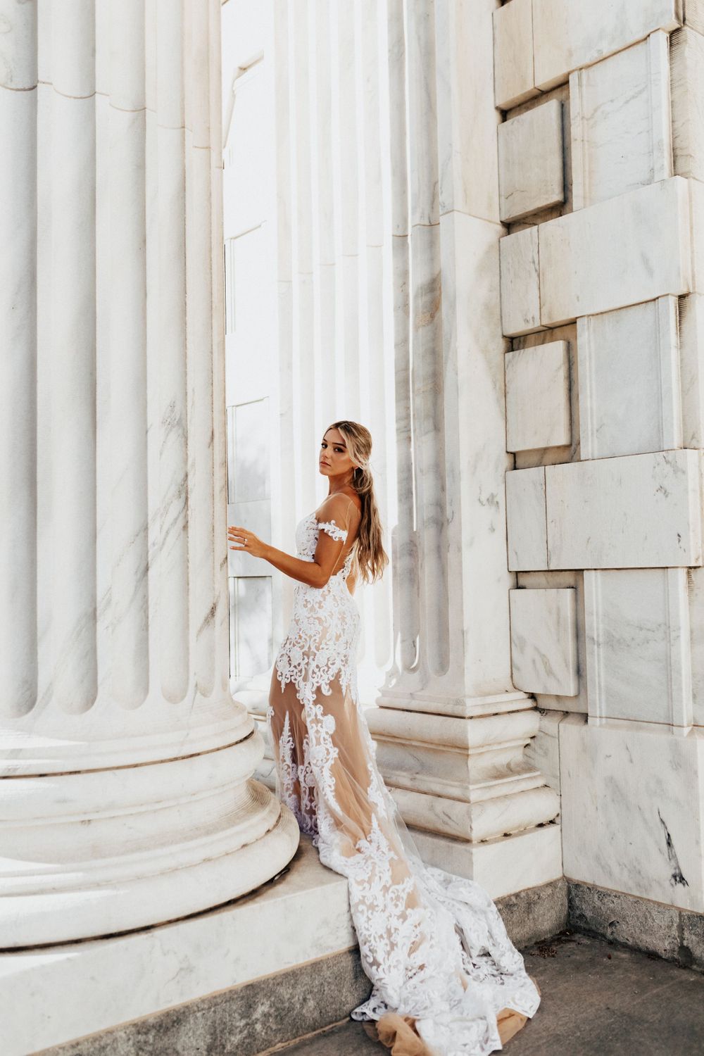 A bride in white lace poses elegantly between classic marble columns.