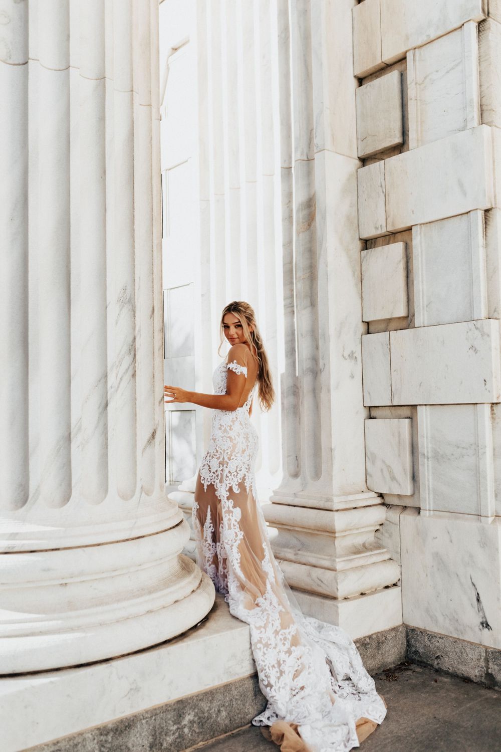 A bride in an intricate lace dress poses between grand white marble columns.