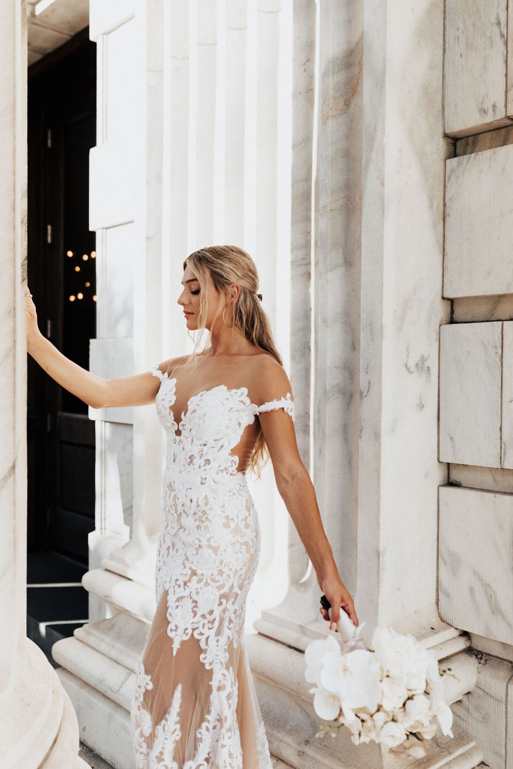 A bride holds white flowers while posing against architectural columns in her lace gown.