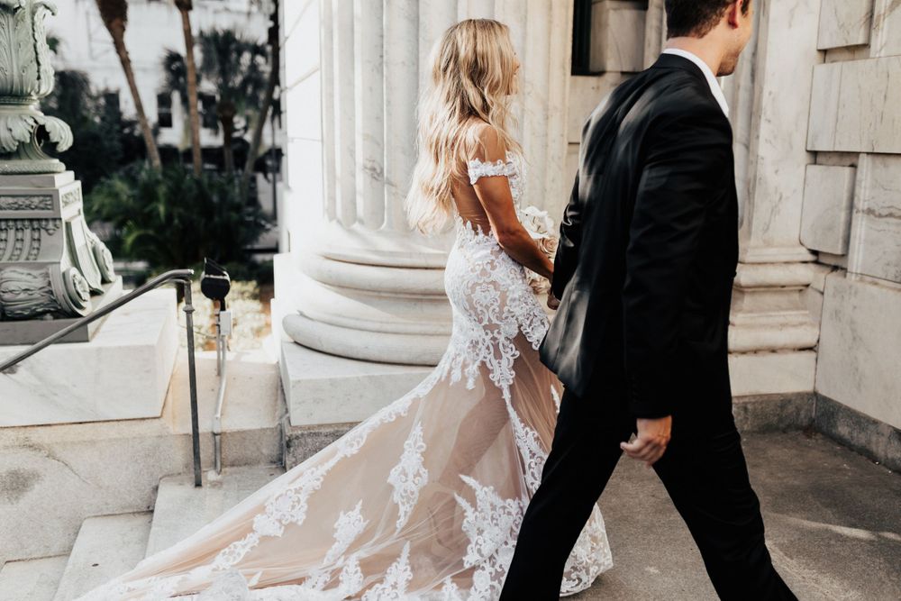 A bride in sheer lace gown walks with groom past elegant white columns.