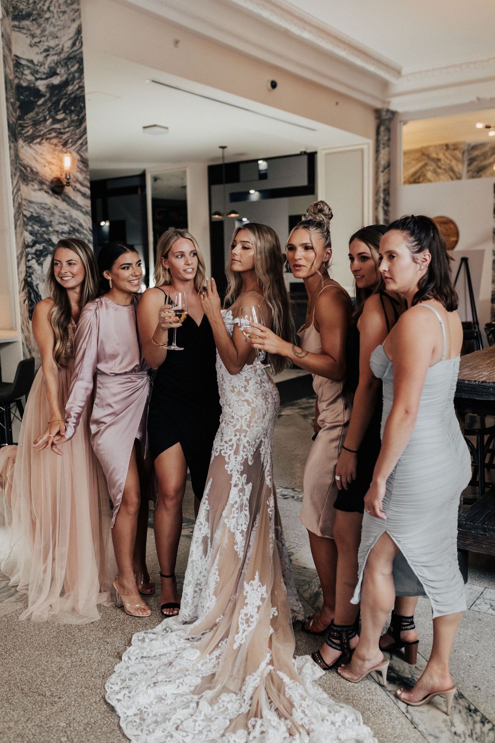 A group of women in formal dresses gather for photos at an elegant indoor venue.