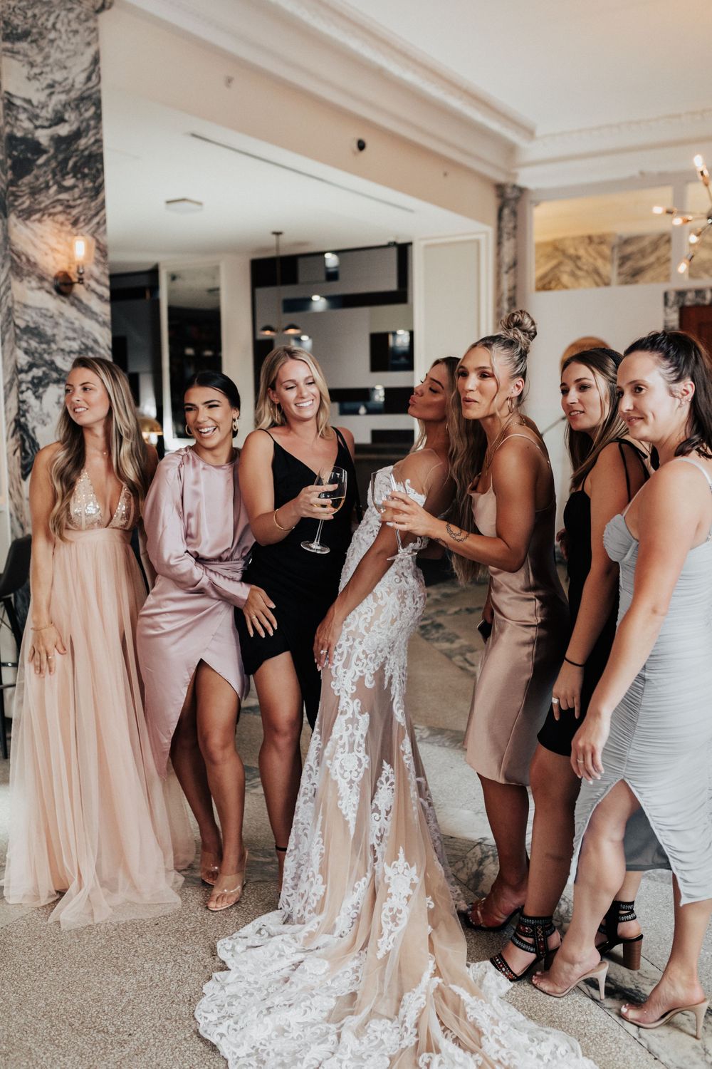 Wedding party in pastel and neutral dresses pose together at an elegant indoor reception.