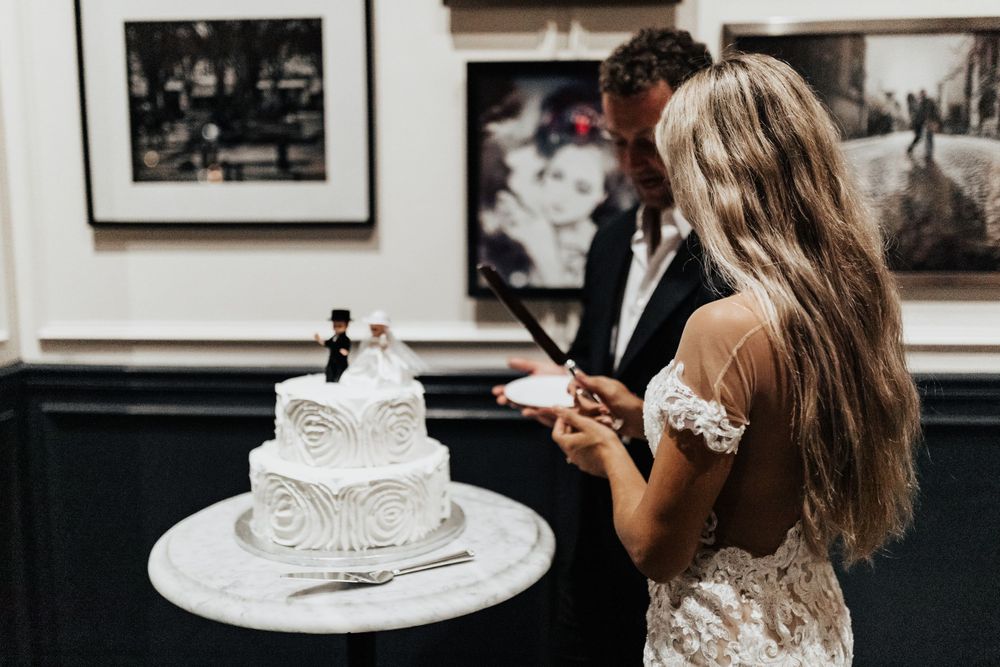 A couple looks at their elegantly decorated white wedding cake in a restaurant with framed photos.