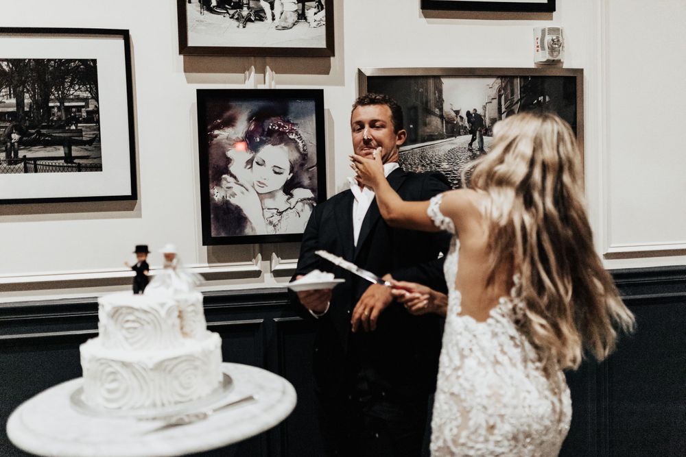 A couple stands beside their white wedding cake while sharing a playful moment.