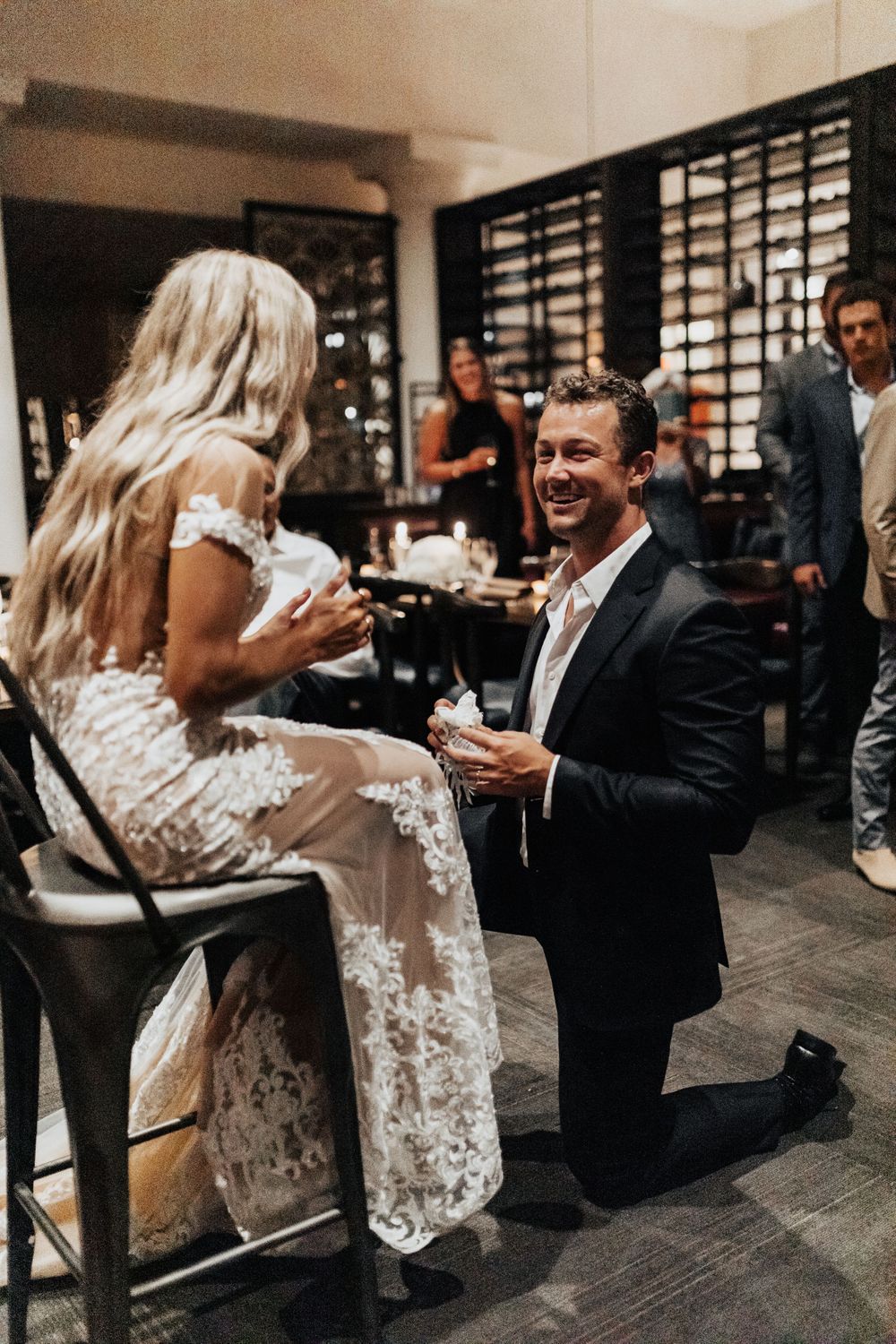 A romantic proposal moment captured in a restaurant with onlookers in the background.