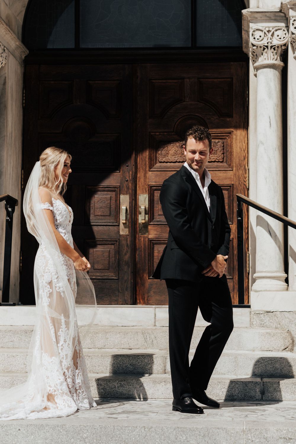 A stylish wedding couple poses against ornate wooden doors and stone columns.