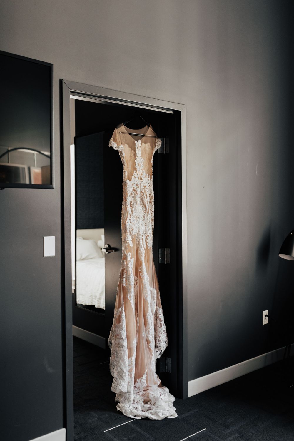 Elegant lace wedding gown hanging in doorway against dark walls in minimalist setting.