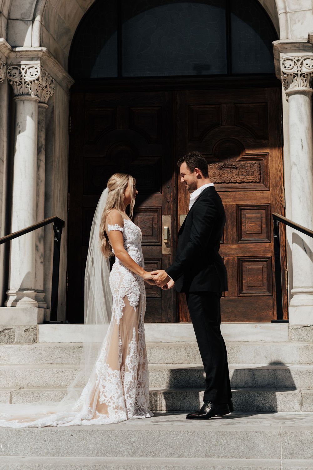 A tender wedding moment captured on church steps with a bride in lace gown and groom in black suit.