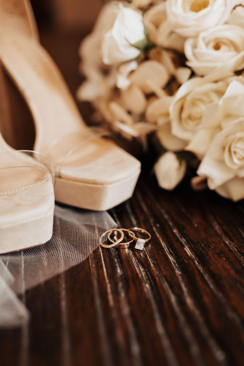 Wedding rings and white shoes artistically arranged with cream roses on a wooden surface.