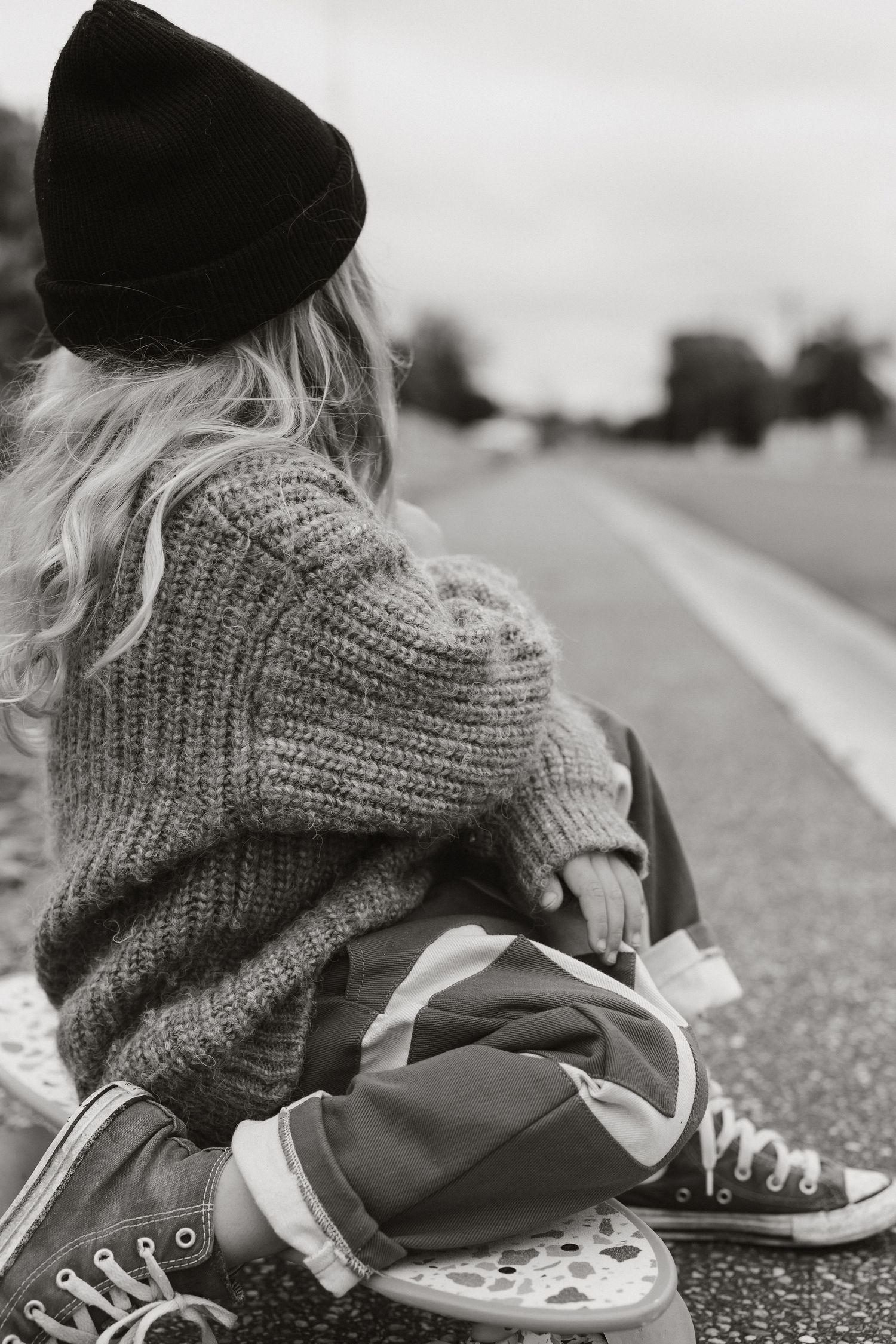 Person in black beanie and cozy sweater sitting on pavement in black and white photography.