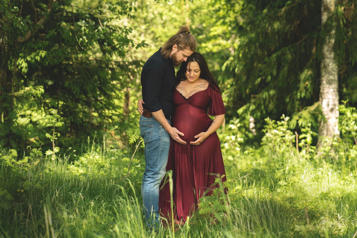 En man håller om sin gravida kvinna där de tittar kärleksfullt ner på kvinnans gravidmage. En solig sommardag.