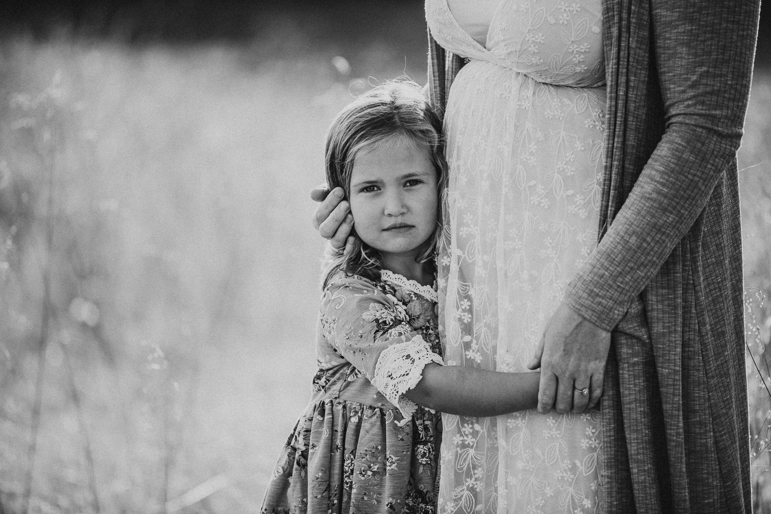Black and white portrait of child holding parent's dress in dreamy field setting.
