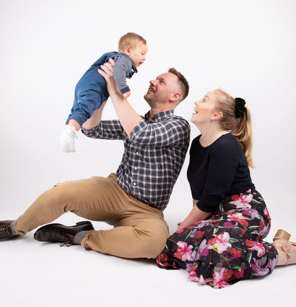 Family Photo Shoot within Solihull studio with toddler and his parents. Little boy is being held in the air by dad, as mom watches lovingly