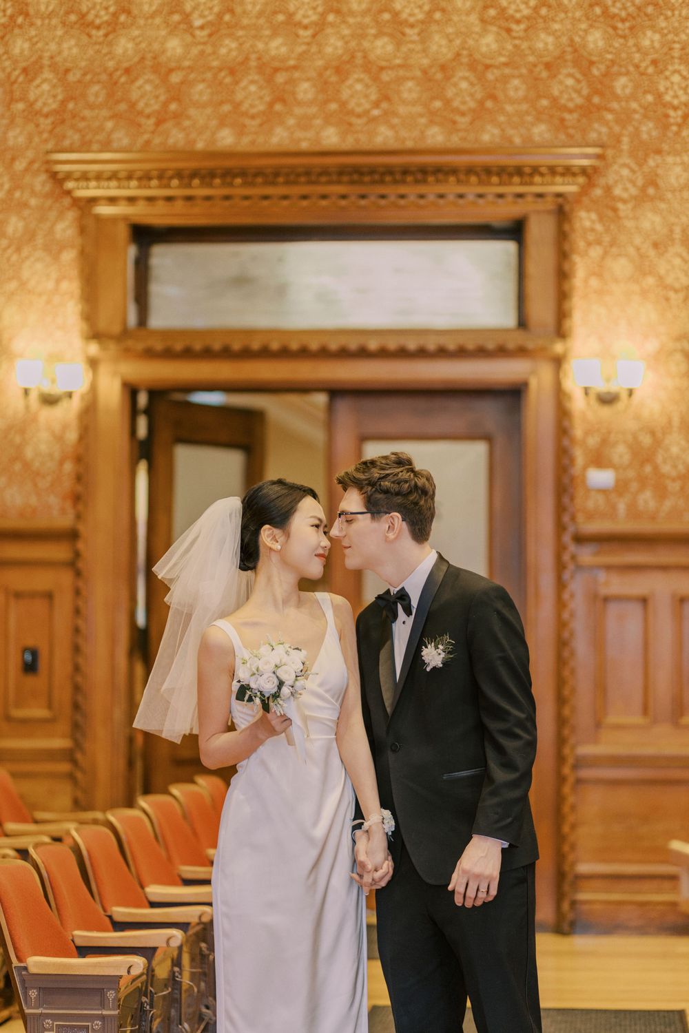 A tender moment between newlyweds in an ornate room with orange wallpaper and wooden details.