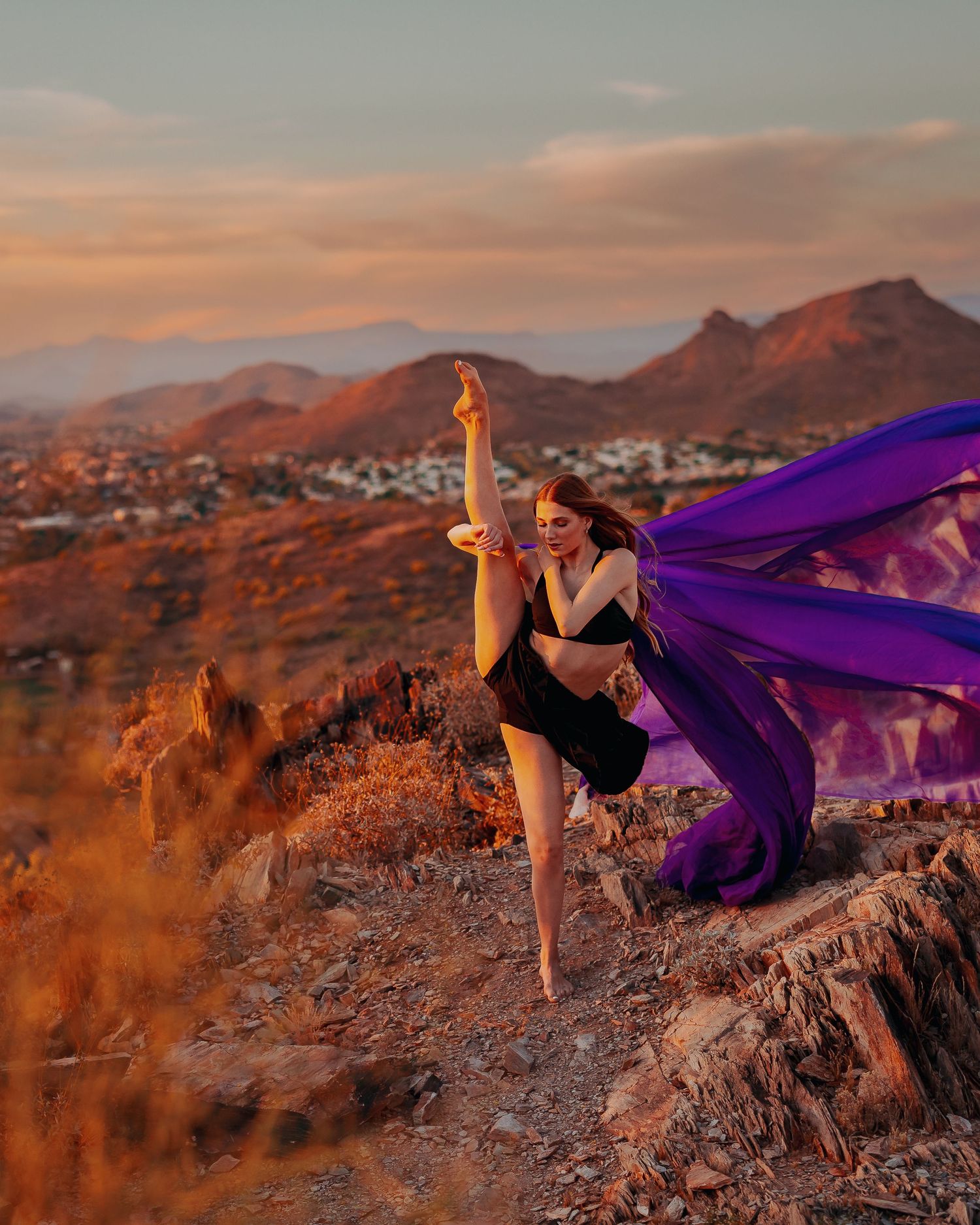 Dancer strikes dramatic pose with flowing purple fabric against desert mountain landscape at sunset.