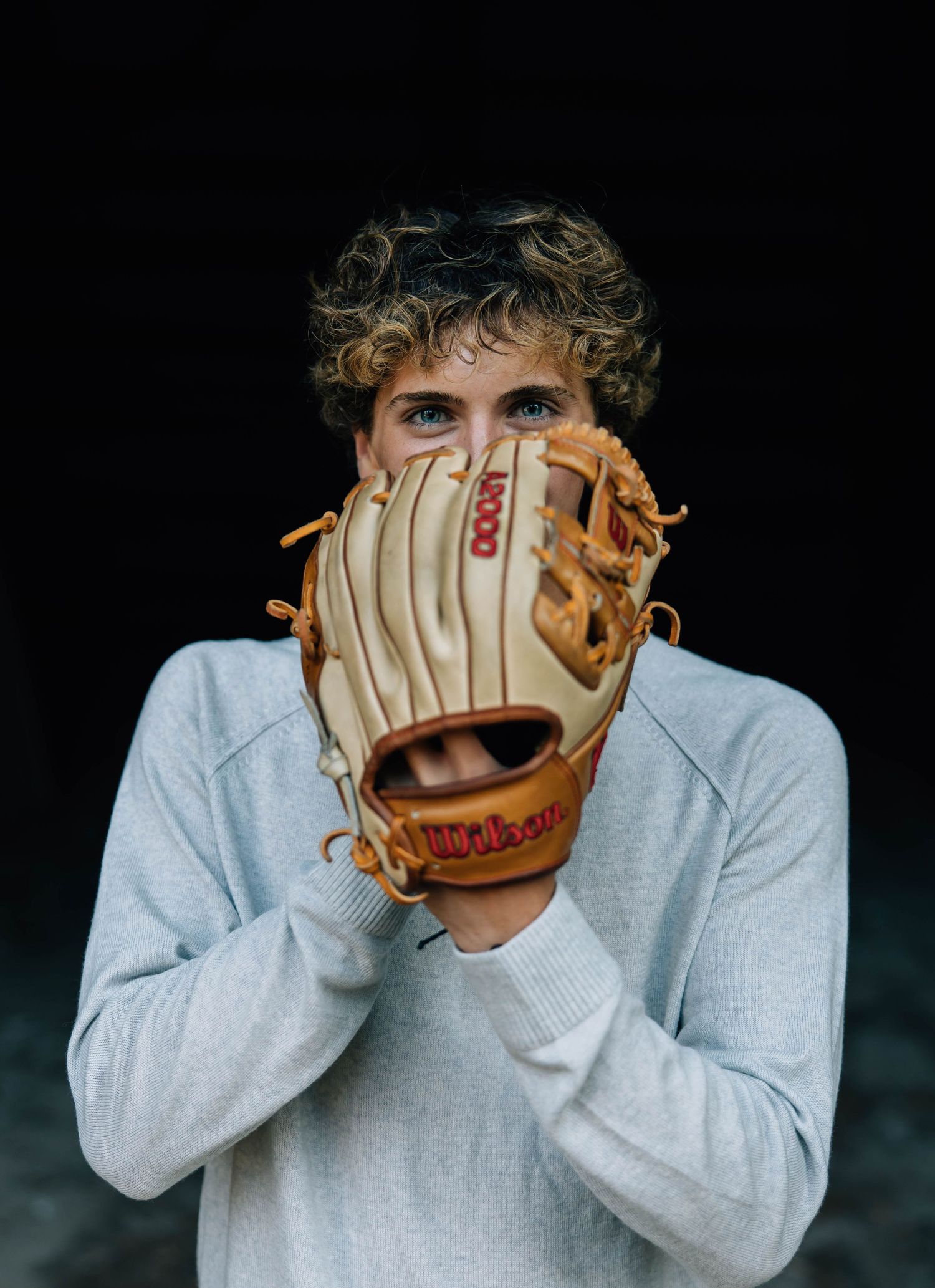 Curly-haired teen holds Wilson baseball glove up to face against dark background.