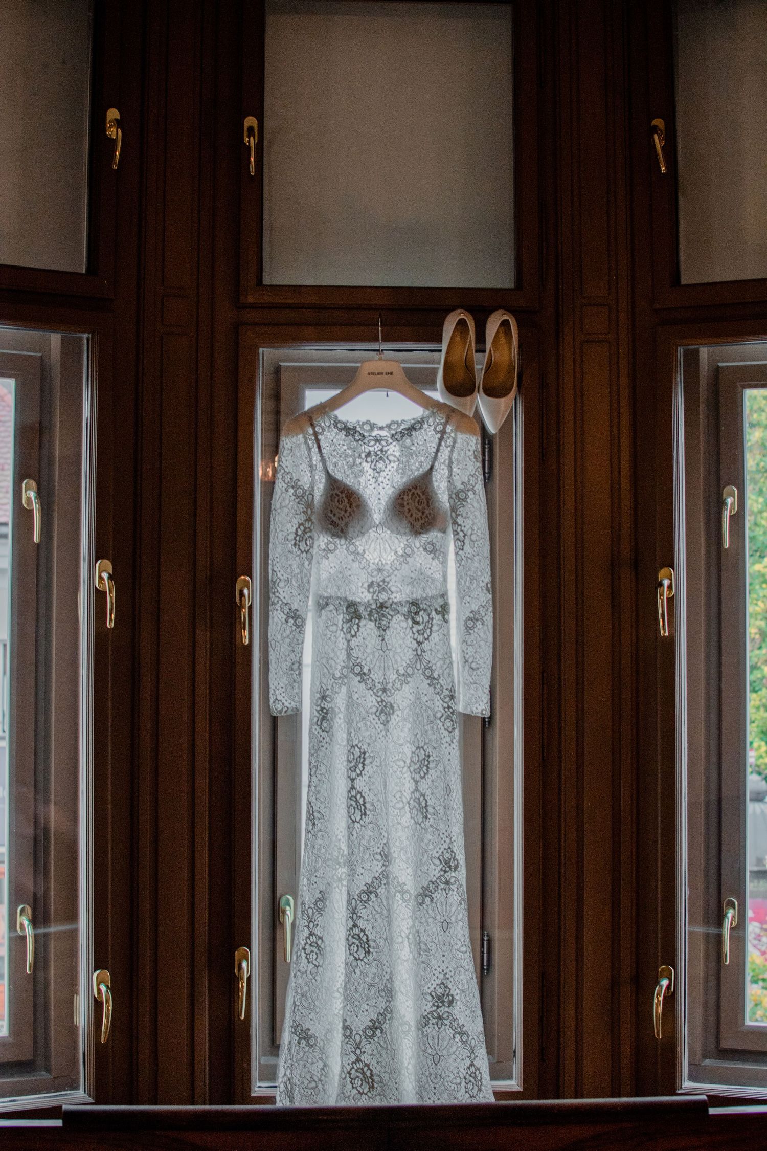 White lace wedding dress and beige heels hanging against dark wooden windows.