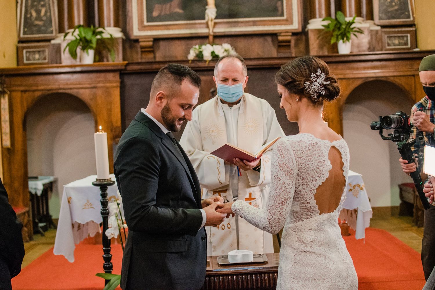 A wedding ceremony takes place in a church with a masked officiant and couple exchanging rings at the altar.