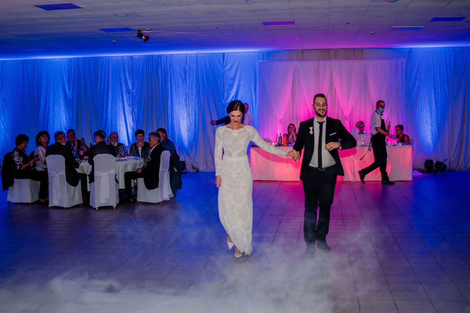 Couple dancing in a reception hall with dramatic blue and pink lighting and fog effects.