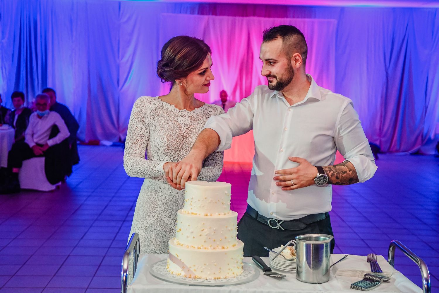 Couple cutting their elegant white wedding cake together at their reception with dramatic pink and blue lighting.