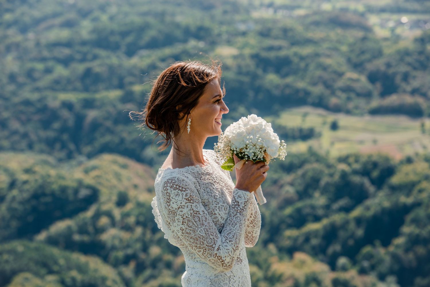 Bride in lace dress holds white bouquet while smiling against mountain landscape backdrop.