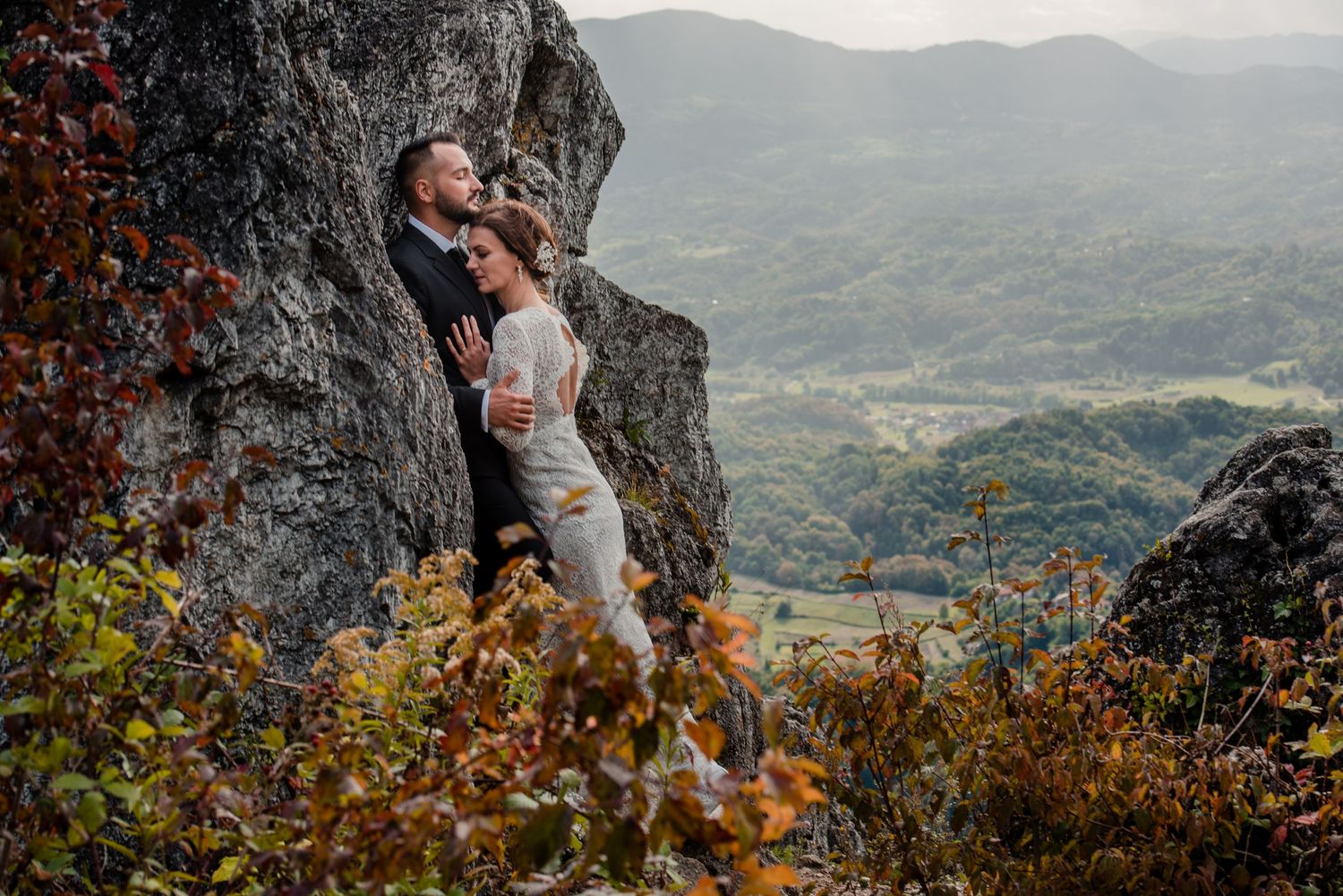 Wedding couple embracing on dramatic mountain cliff with scenic valley views.