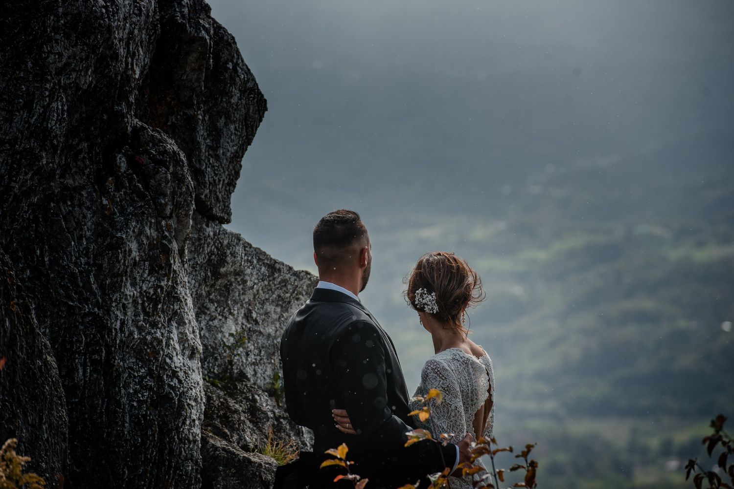 Wedding couple stands on mountain overlook against moody sky and distant hills.