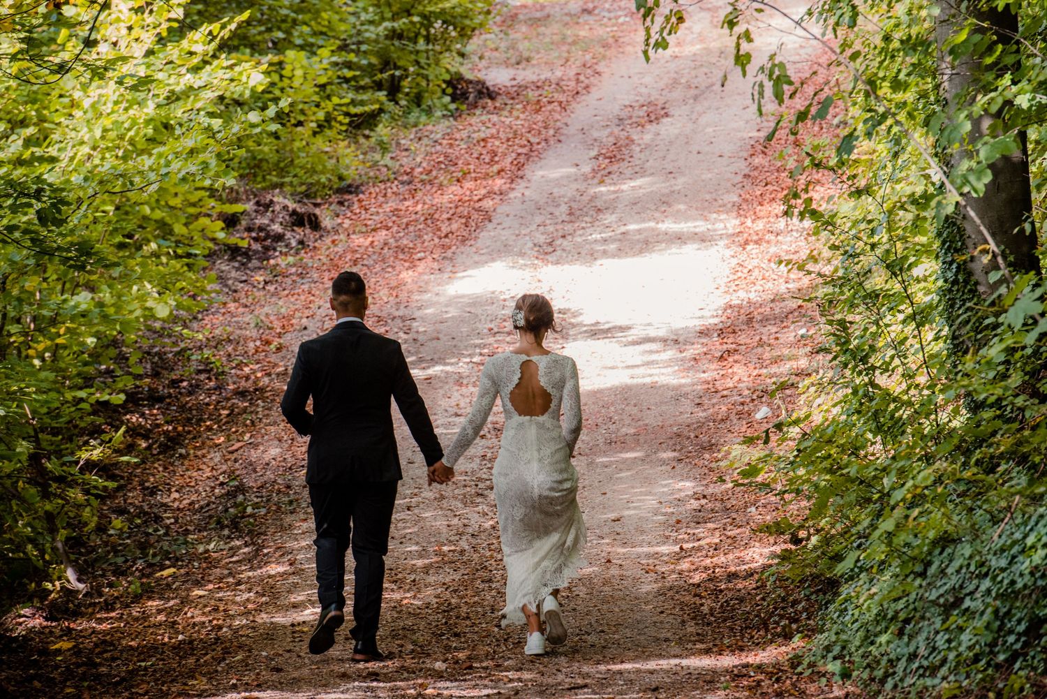 Newlyweds walking hand in hand down tree-lined path in elegant wedding attire.