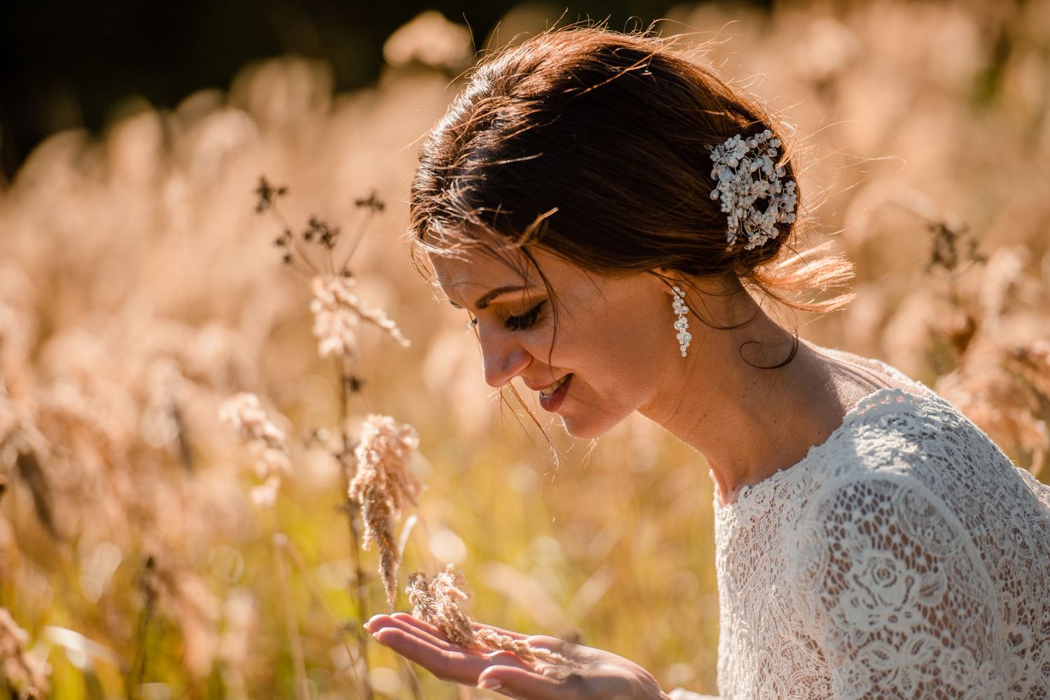Bride in lace dress holds dried flowers in golden sunlit field.