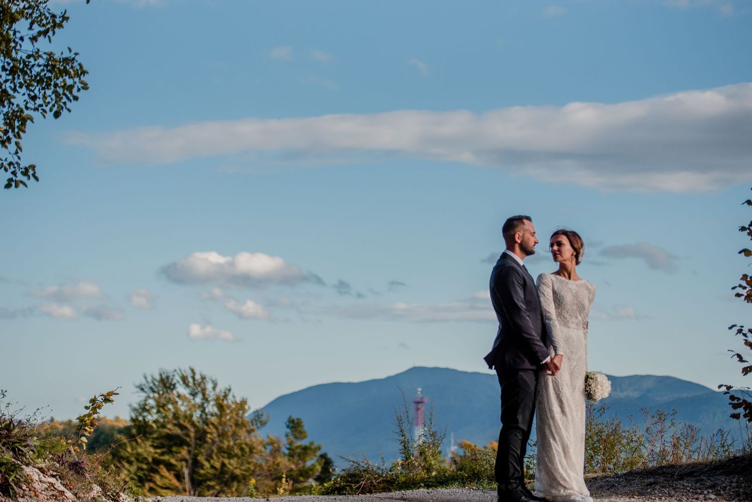 A couple stands together against a mountain landscape backdrop with blue sky and puffy clouds.