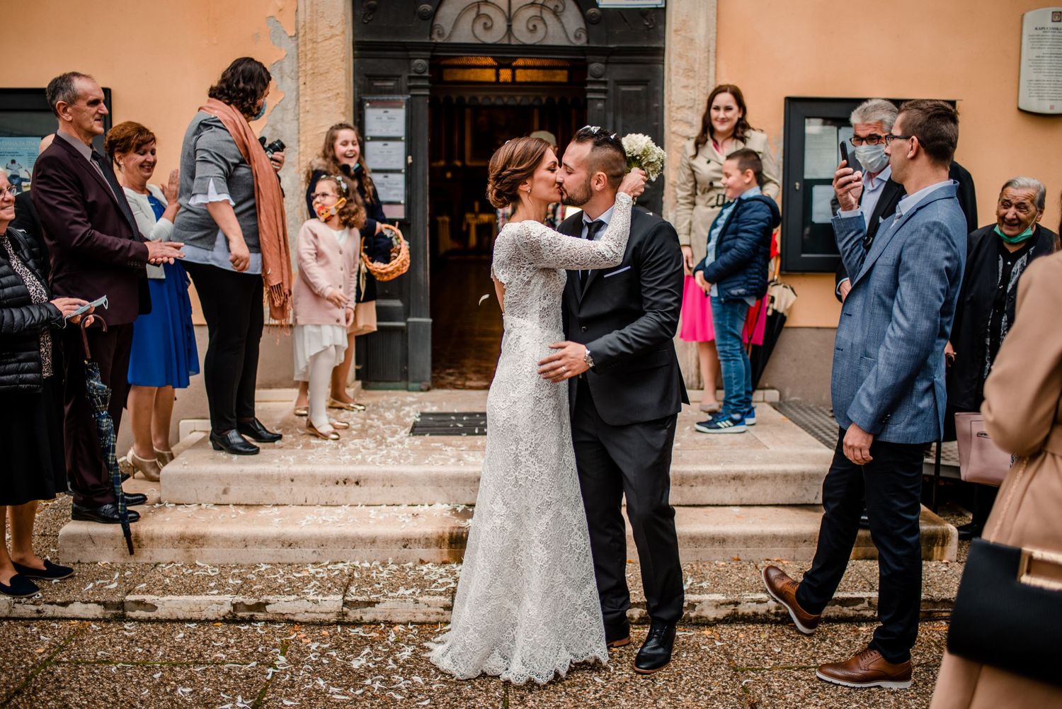 Wedding celebration outside a historic building with gathered guests watching the newlyweds share a moment.