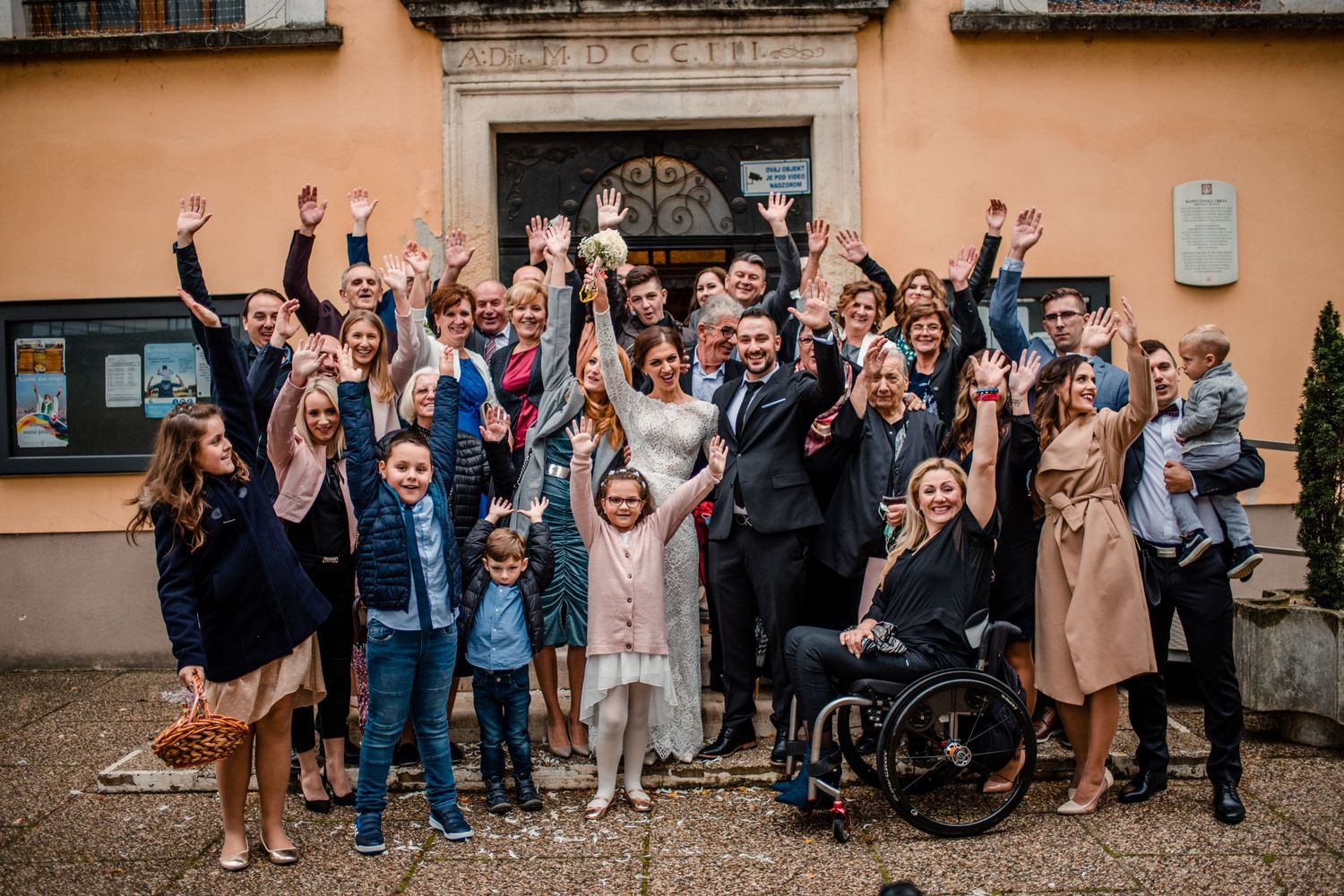 Large wedding group photo with guests celebrating outside a peach-colored historic building.