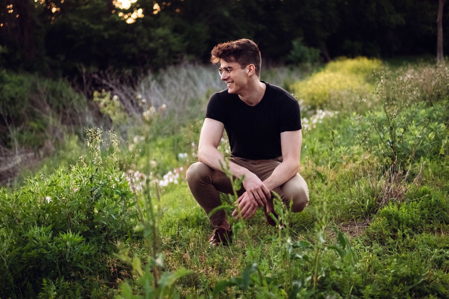 A person in a black t-shirt crouches in a lush green meadow at sunset.