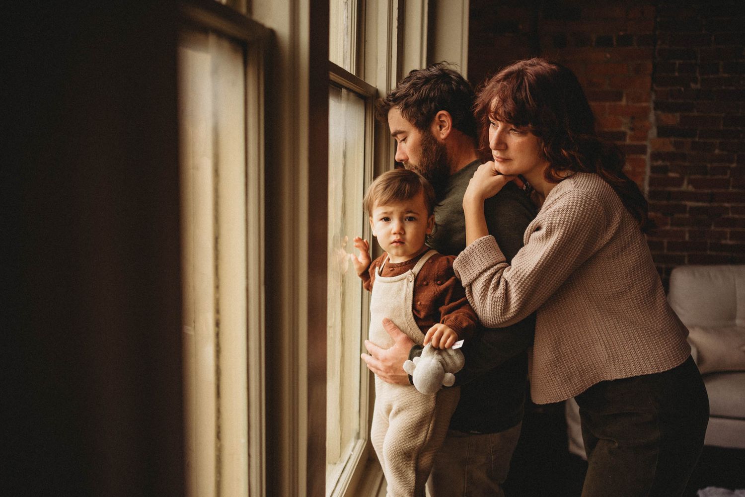 Family gazes through large windows together in a brick-walled room.